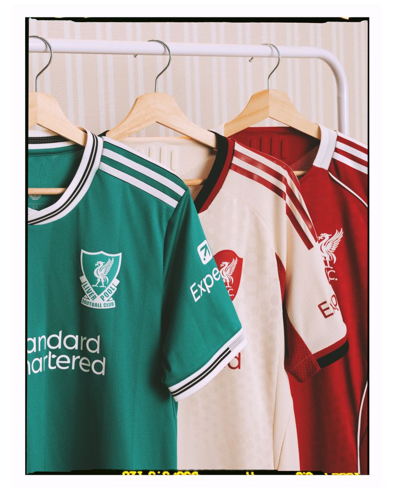 Three soccer jerseys hang on wooden hangers from a clothing rack, featuring the Liverpool Football Club logo and an off-white striped background.