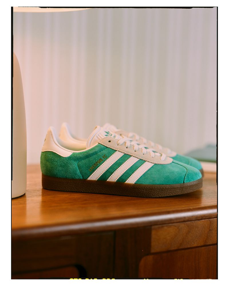 Close-up of green Adidas Gazelle sneakers with white stripes on a wood surface against a light background, highlighting the shoe's design.