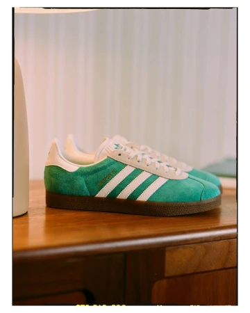 Close-up of green Adidas Gazelle sneakers with white stripes on a wood surface against a light background, highlighting the shoe's design.