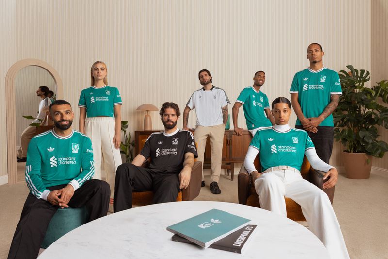 Group of seven diverse adults in a bright interior setting, wearing soccer jerseys, sitting and standing around a round table.