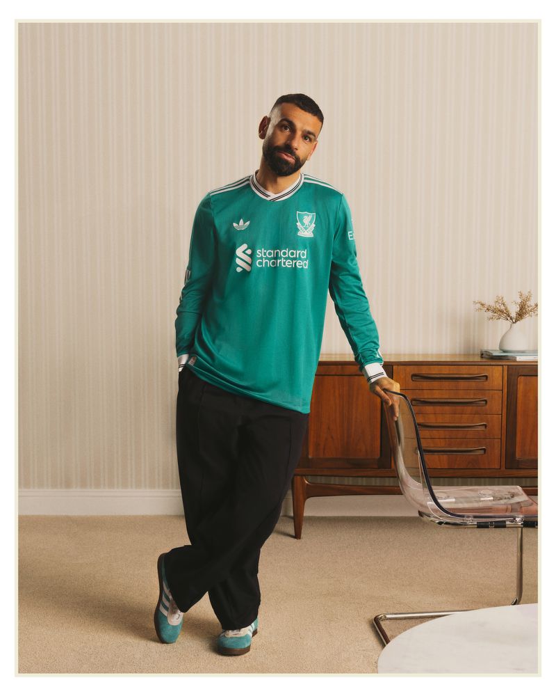 Man in a green Liverpool soccer jersey and black pants posing in an indoor environment with a clear chair and wood cabinet.