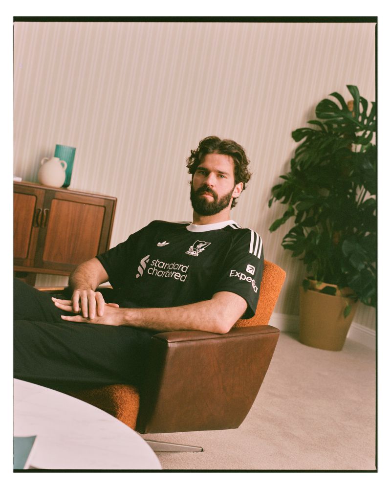 Adult male in a black Liverpool soccer jersey poses in an orange chair in a room with a mid-century modern design and a houseplant.