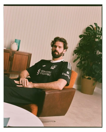 Adult male in a black Liverpool soccer jersey poses in an orange chair in a room with a mid-century modern design and a houseplant.