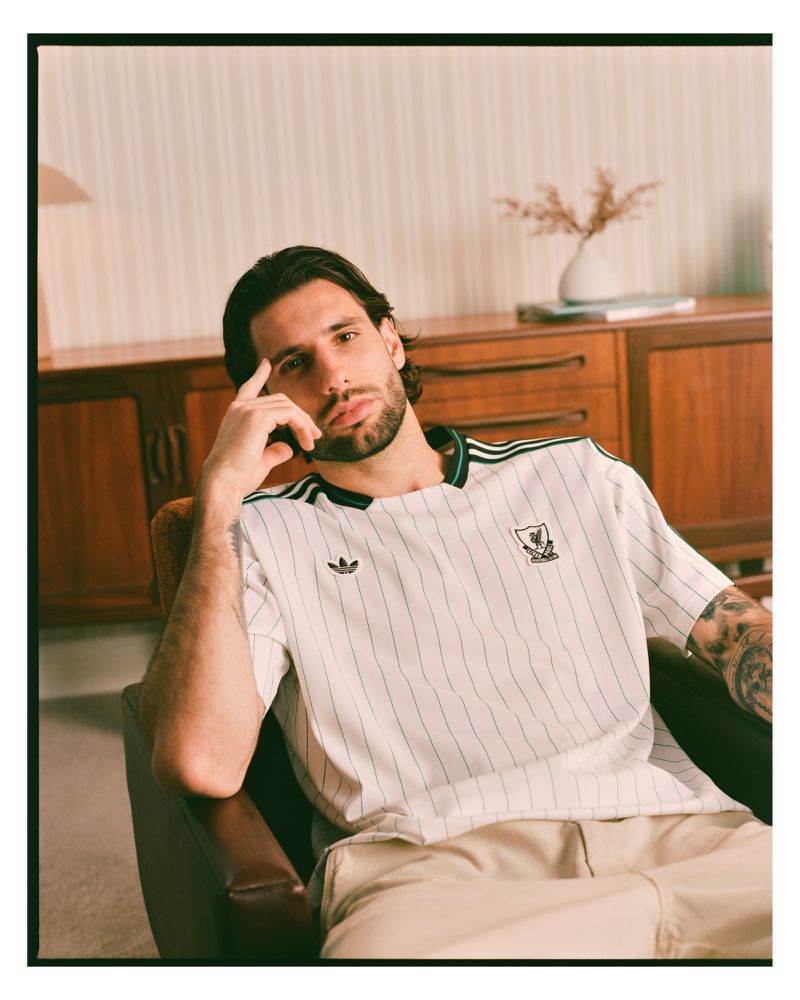 Portrait of an adult male in a sports jersey sitting in a chair inside a living room with minimalist decor, styled for an Adidas ad.