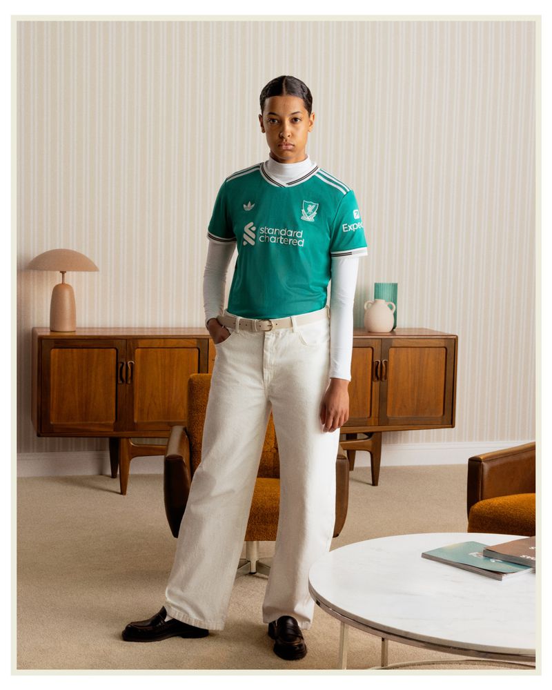 Young adult in a teal soccer jersey stands in a mid-century modern living room with vintage furniture and a beige striped wallpaper backdrop.