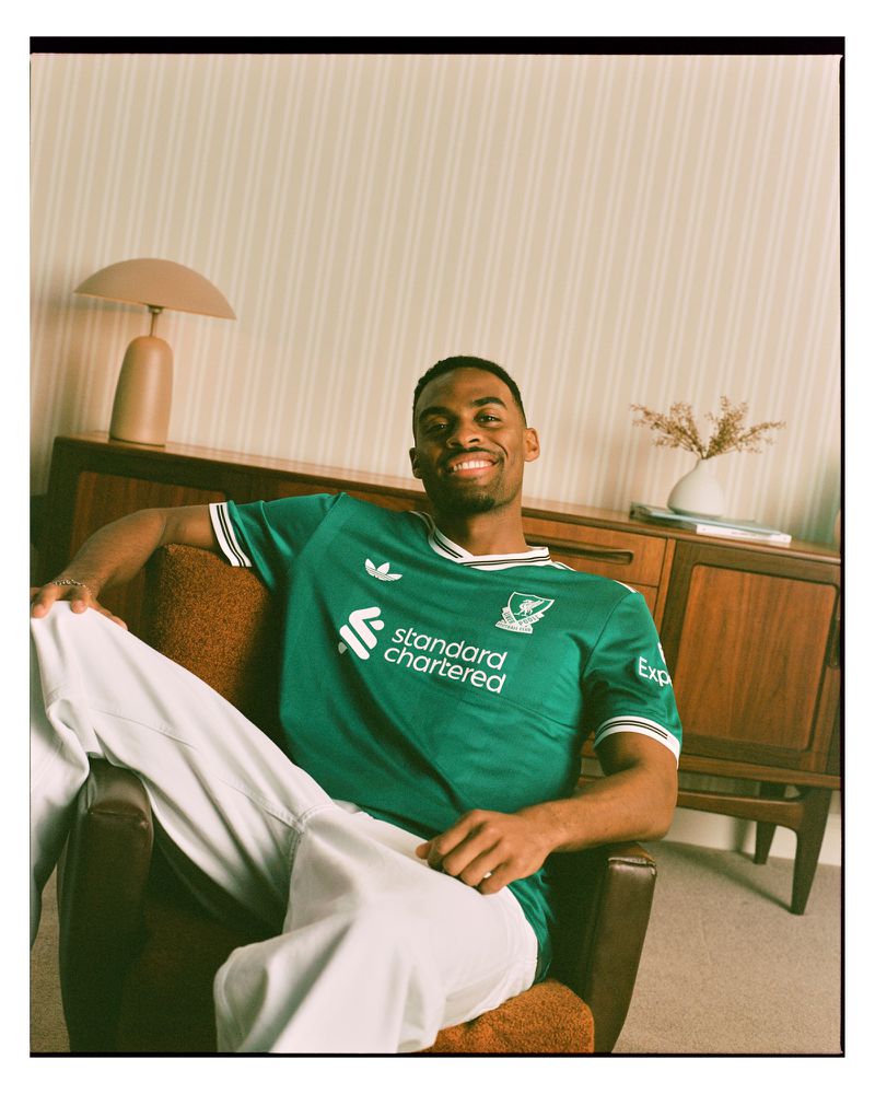 A smiling Black man in a green soccer jersey poses in a chair in a warm-toned interior with retro-style furniture and natural light.