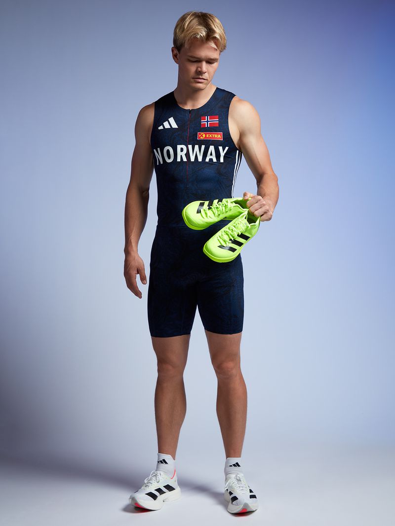 Athletic man in sports attire stands holding neon yellow running shoes in studio setting with gradient blue to white background.