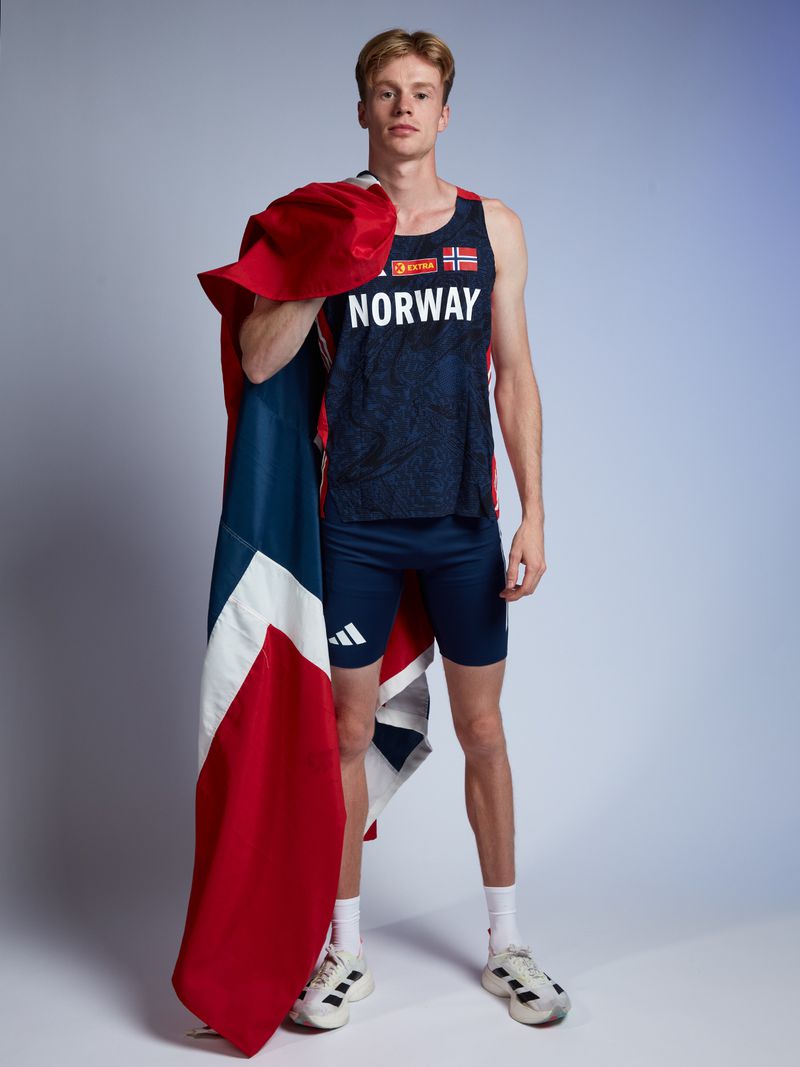 Male athlete poses in athletic wear indoors against a gradient background, draped in the Norwegian flag. Sports and national pride are themes.