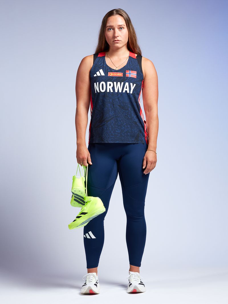 Full-length portrait of an athletic woman in sportswear holding neon yellow boxing shoes, standing against a light blue backdrop.