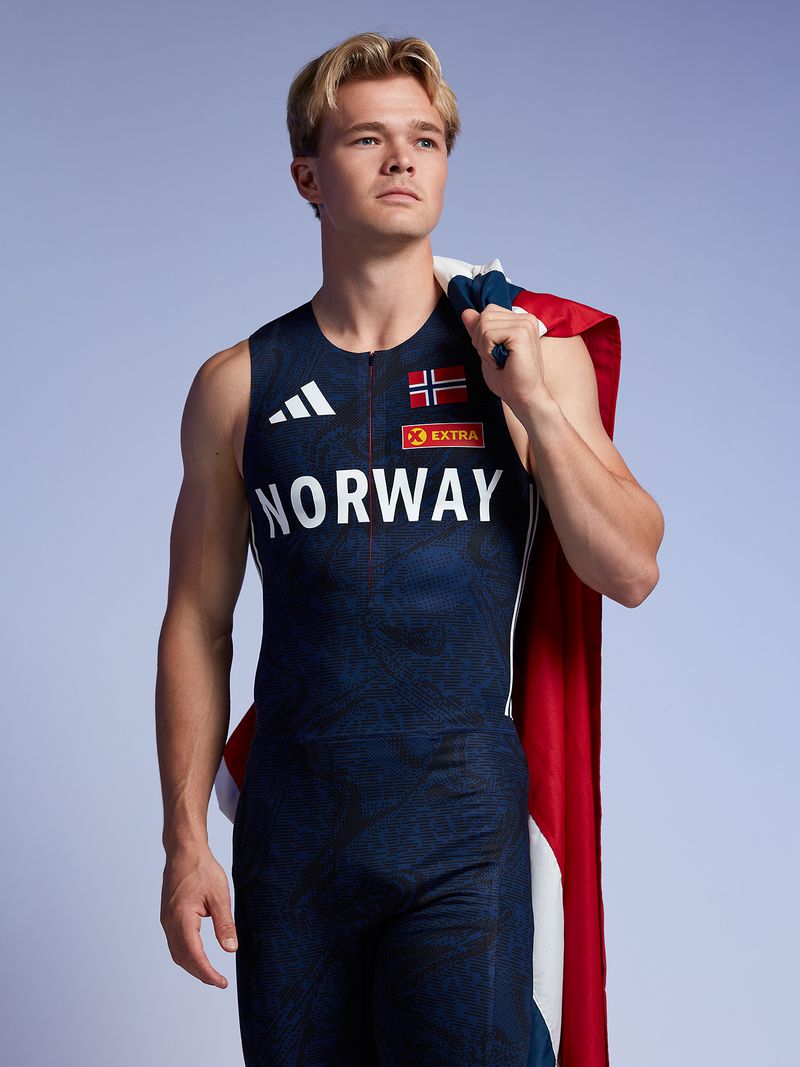 Blond male athlete in blue Norway uniform with the Norway flag draped over his shoulder, posing against a gradient blue background.