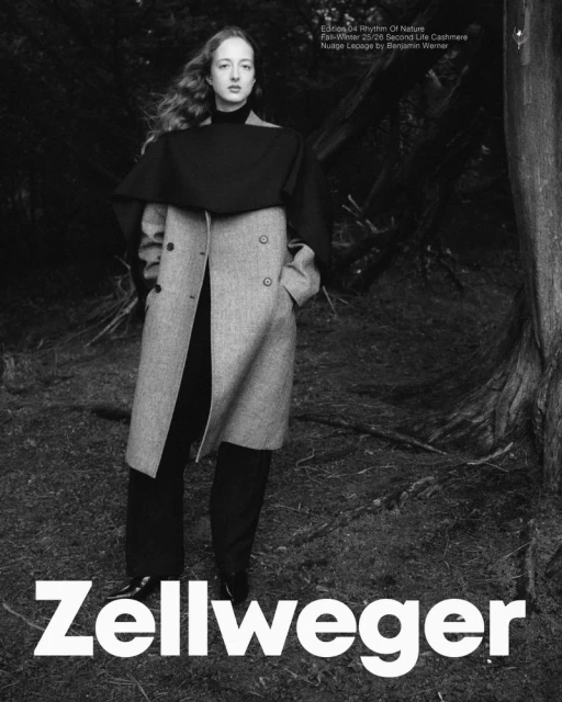 Black and white fashion portrait of a woman with long hair wearing a gray coat and standing in a wooded, outdoor setting.