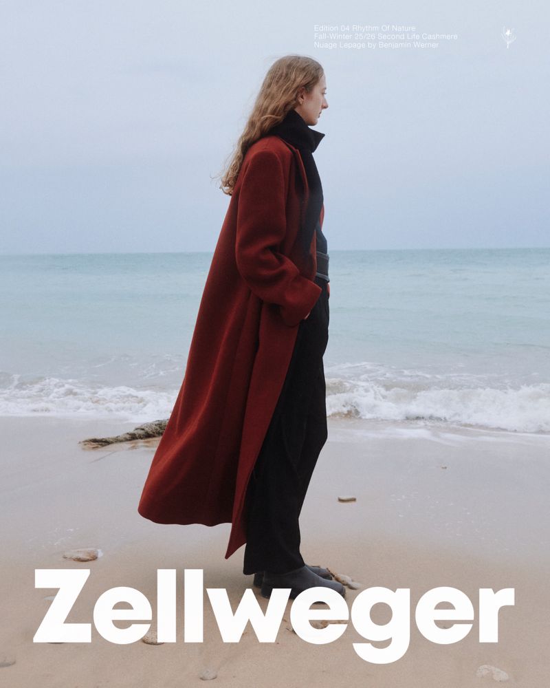 A slender woman in a long red coat poses on a beach with the ocean and sky creating a serene backdrop on a cloudy day.