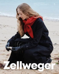 Model kneeling on a sandy beach wearing a dark coat, red scarf, and black leather gloves for Zellweger