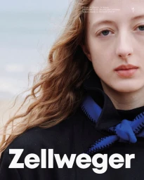 Close up portrait of a young female model with long wavy hair wearing a black jacket and blue scarf against an ocean background.
