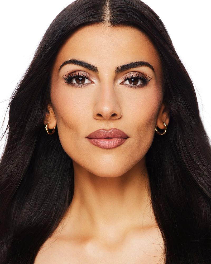 Close-up portrait of a beautiful woman with long dark hair, gold hoop earrings, and light pink lipstick against a white background.