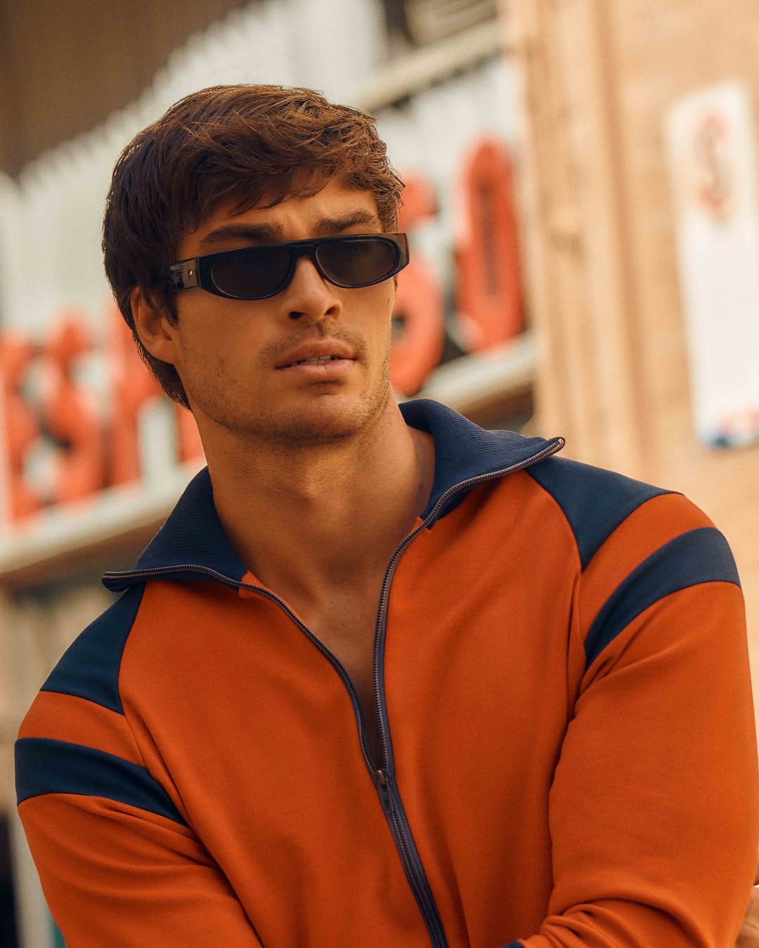 Close-up of an adult male wearing sunglasses and an orange and navy blue jacket, posing outdoors during daytime.