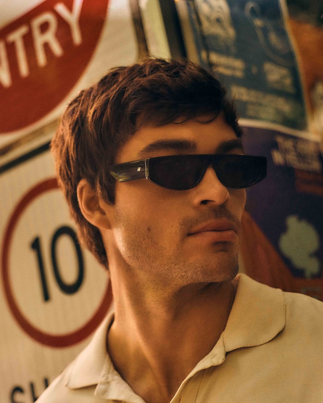 Close-up of a young adult man wearing dark sunglasses and a tan shirt, posing in front of blurred signage. Indoor setting.