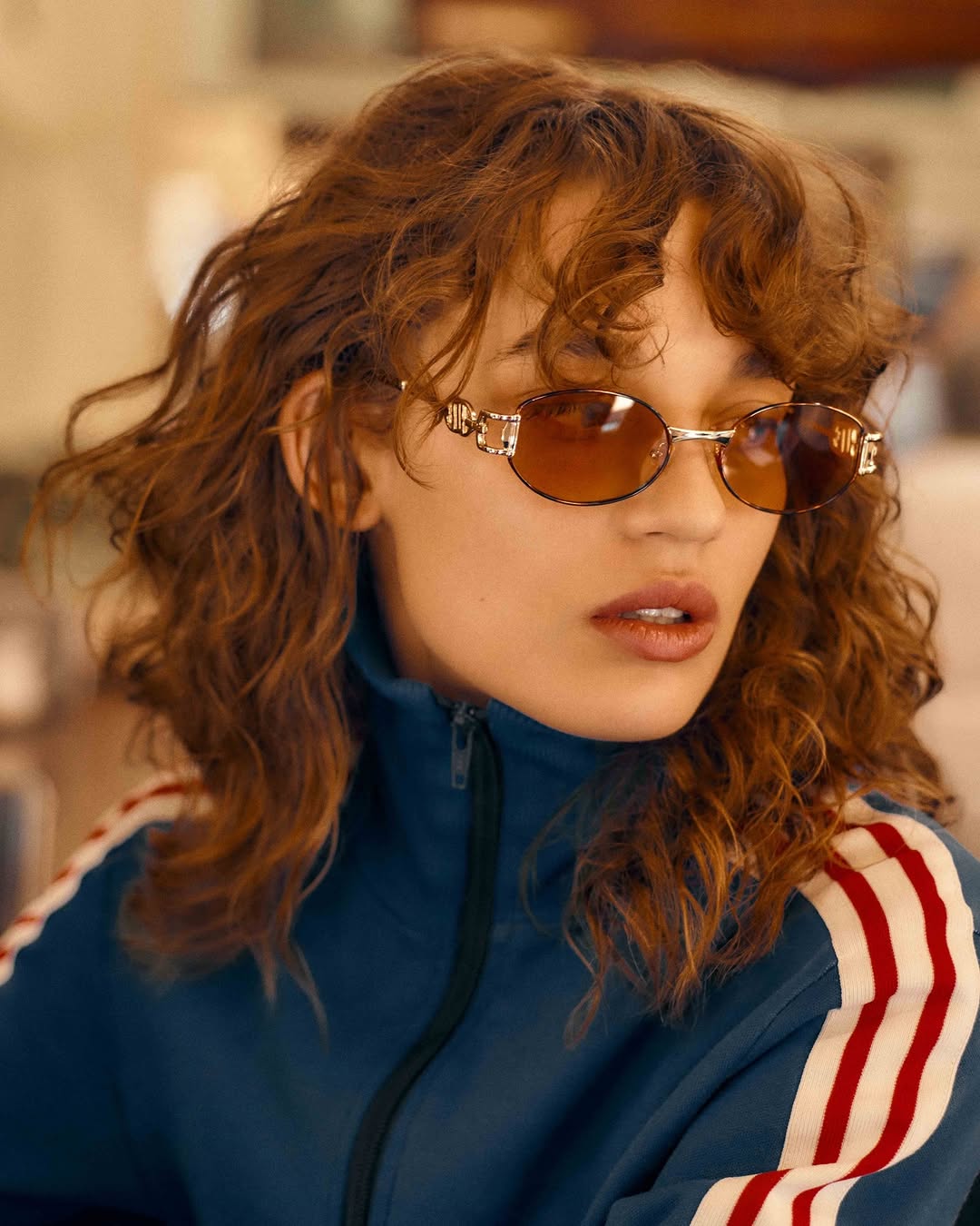 Woman wearing sunglasses and a blue track jacket with white stripes poses indoors with a neutral background, looking forward at the camera.