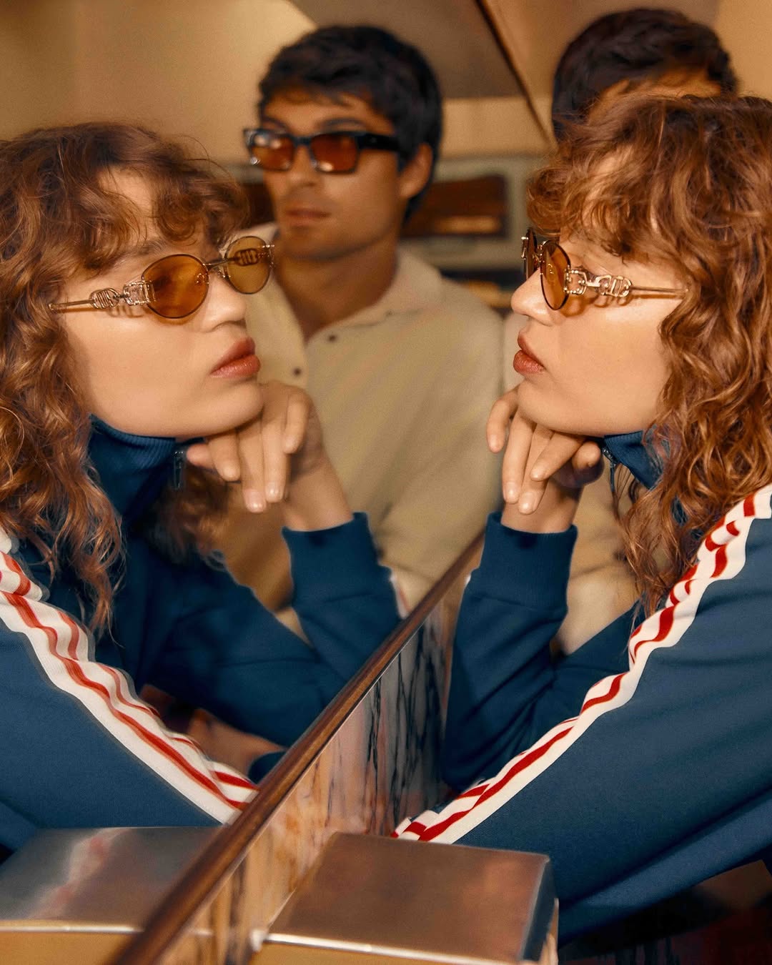 Woman with curly hair wearing sunglasses reflected in a mirror, standing near another person with sunglasses. Fashion and eyewear concept.