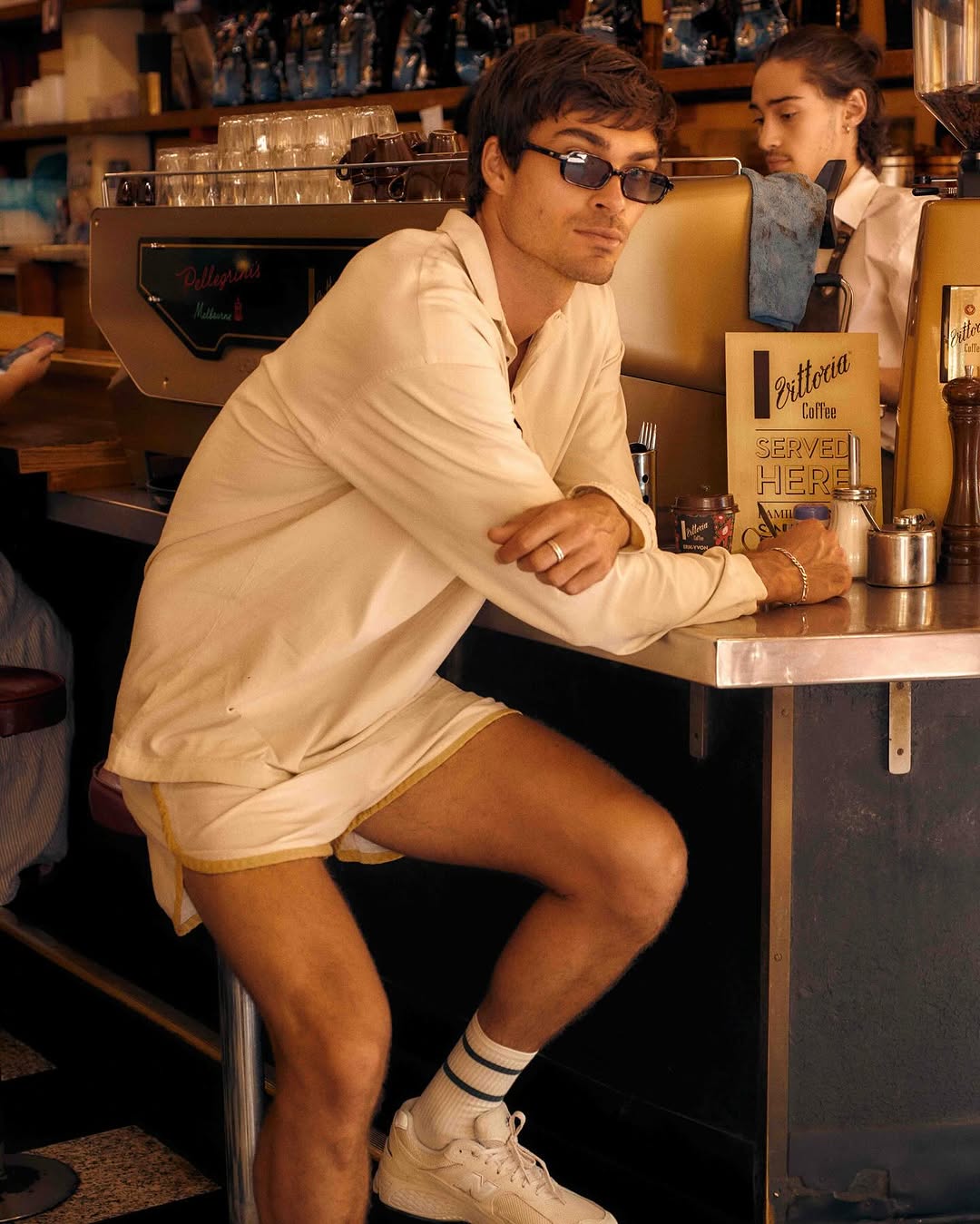 Young man in sunglasses and shorts at coffee shop counter. He is leaning on the counter, wearing tube socks and sneakers.