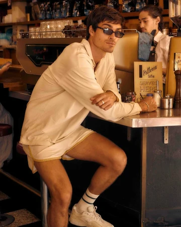 Young man in sunglasses and shorts at coffee shop counter. He is leaning on the counter, wearing tube socks and sneakers.