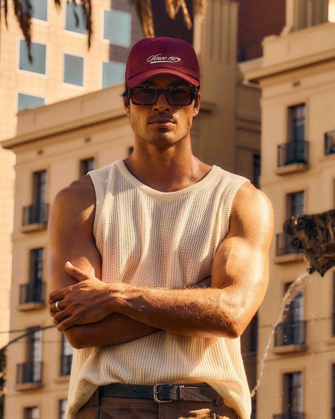 Portrait of a muscular adult male wearing a tank top and baseball cap, posing outdoors in an urban environment during daytime.