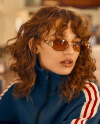 Model with curly brown hair wearing tinted sunglasses and a blue track jacket for Le Specs New Vintage campaign