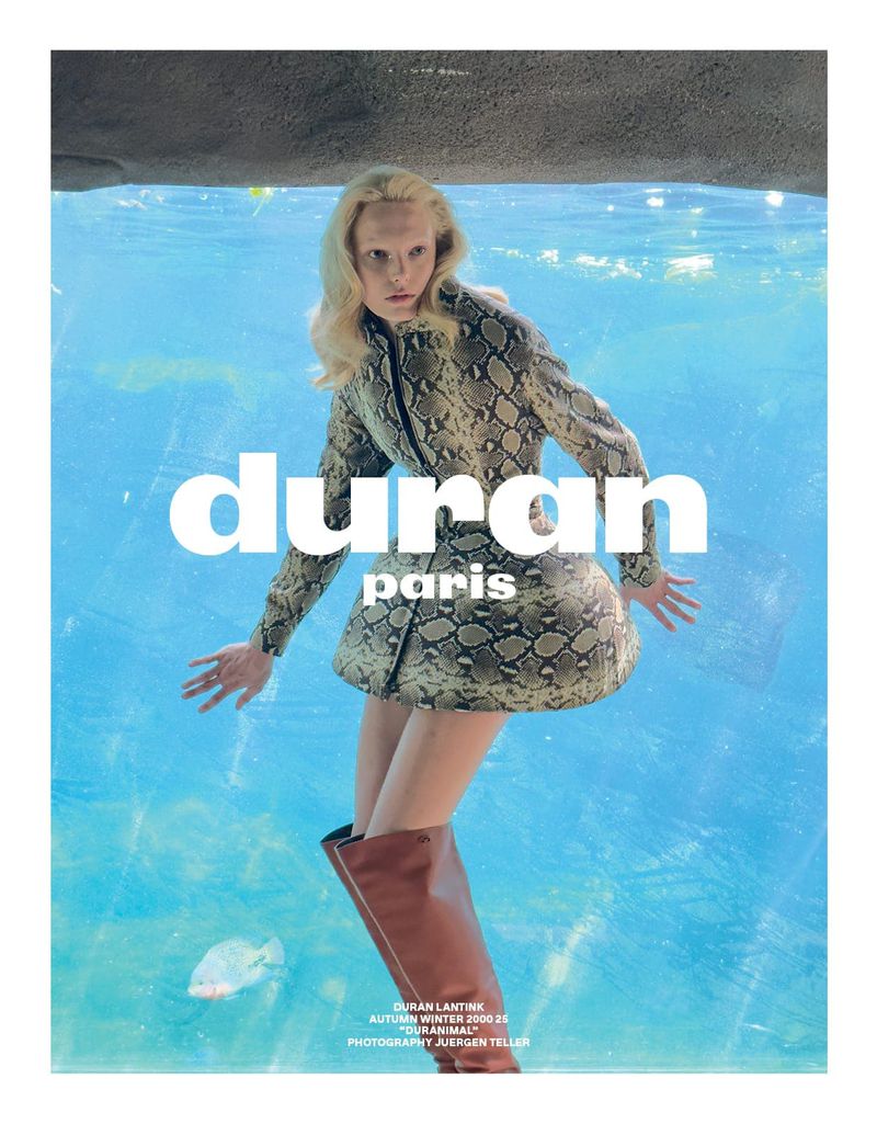 Fashion editorial shot: Woman in snakeskin-patterned suit and brown boots, posing underwater in a tank with a fish.