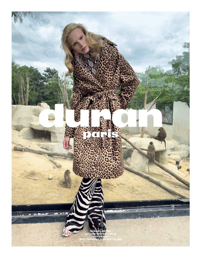 Fashion editorial image featuring a fair-skinned woman in an animal-print coat and zebra-striped pants, posing in front of monkeys.