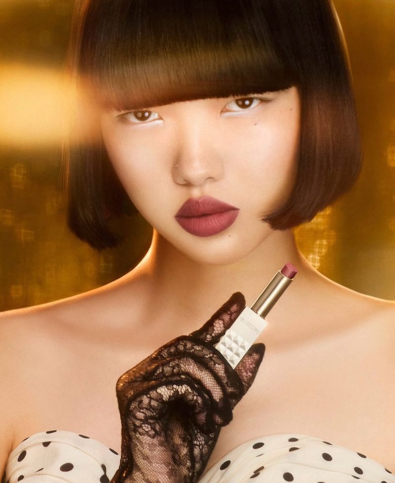 Asian woman in studio, wearing lipstick and lace gloves, holding a lipstick tube, gold background with dark brown bob haircut.