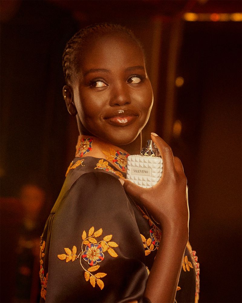 Dark-skinned woman holds Valentino Born in Roma Donna perfume bottle, black floral-embroidered robe, warm studio lighting, beauty shot.