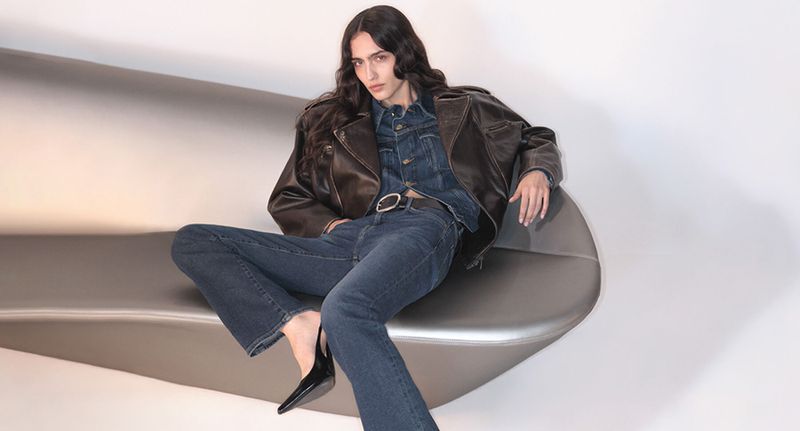 An adult female model with long dark hair poses on a sculptural bench wearing a brown leather jacket, denim shirt, and dark blue jeans.