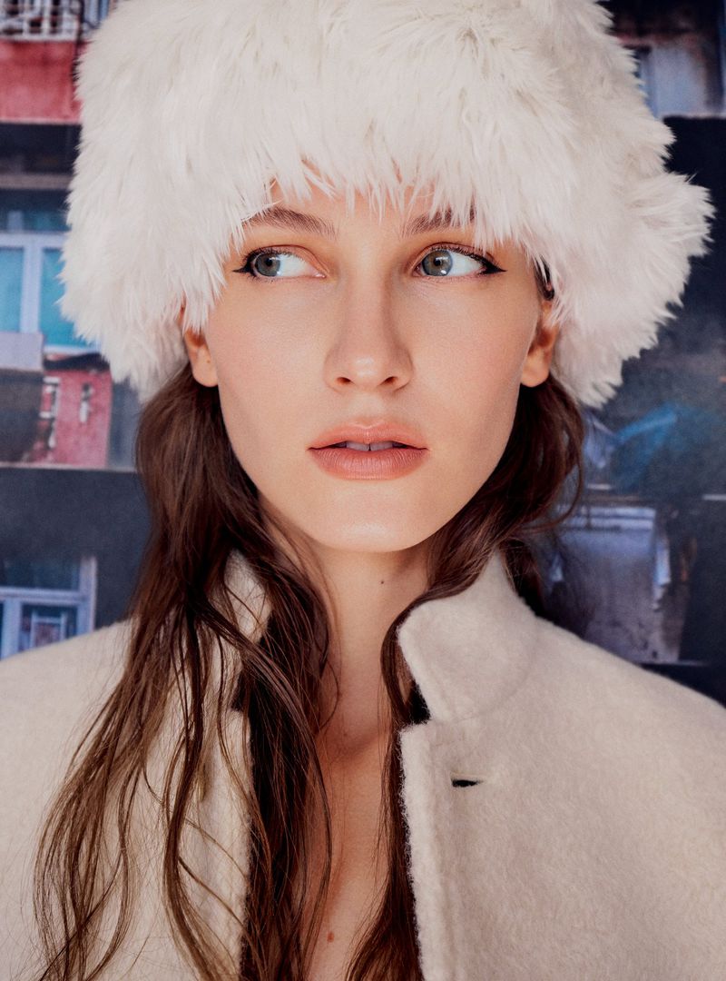 Close-up of a Caucasian woman in a white furry hat and cream-colored coat, looking off to the side against an abstract background.