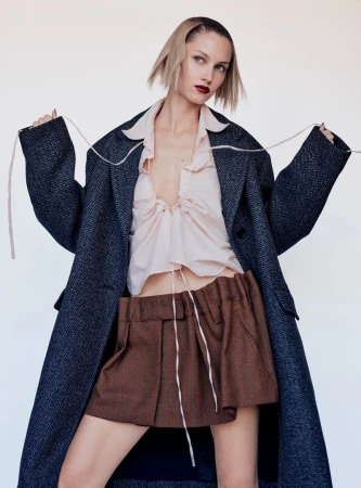 Model Claudia Campana wearing an oversized dark coat, pale ruffled blouse, and brown pleated mini skirt against a white studio background.