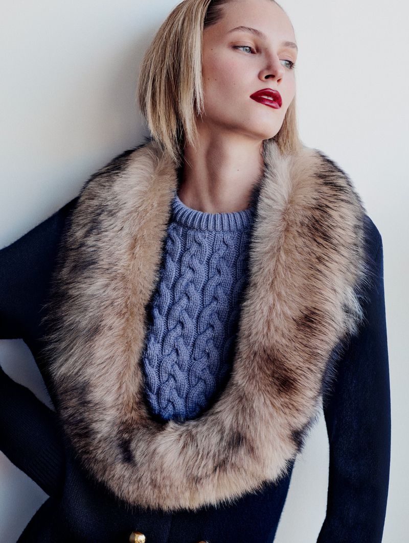 Blonde woman in a blue cable-knit sweater and black coat with a fur collar, posing indoors in front of a white wall.