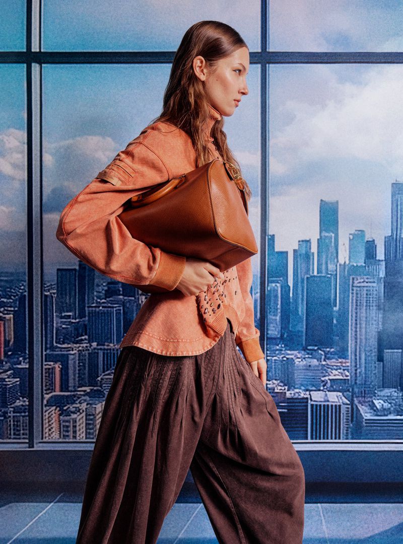 Fashionable adult woman in a salmon-colored jacket and brown pants poses with a brown leather handbag in front of a cityscape backdrop.