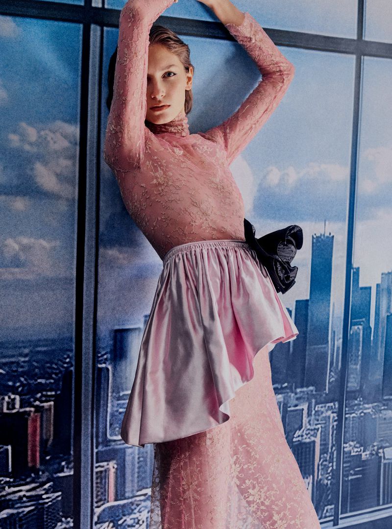 A slender model in a pink lace dress with a dramatic skirt poses in front of a blurred cityscape backdrop, fashion shot, portrait.