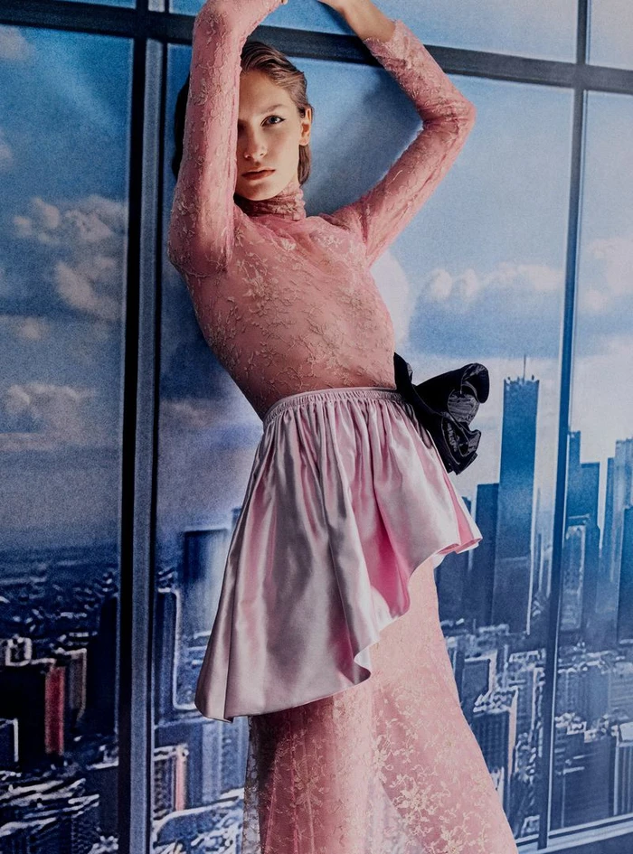 Model in a sheer pink lace dress with a satin peplum poses against a window backdrop overlooking a city skyline.