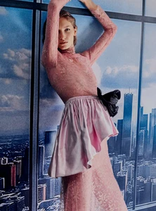 Model in a sheer pink lace dress with a satin peplum poses against a window backdrop overlooking a city skyline.