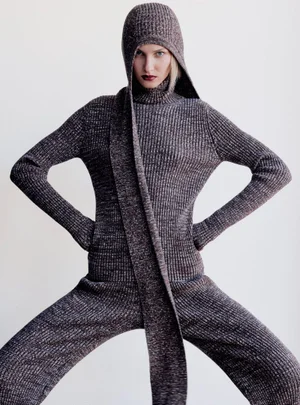 Model wearing a marled ribbed knit hooded jumpsuit and scarf posing with hands on hips against a white studio backdrop.