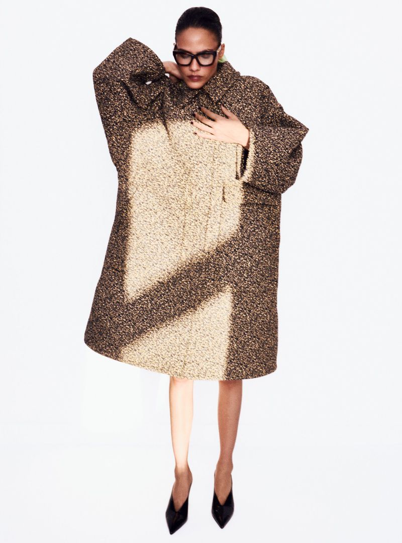 A woman in an oversized, speckled coat and black heels poses in a brightly lit studio. The focus is on fashion and style.