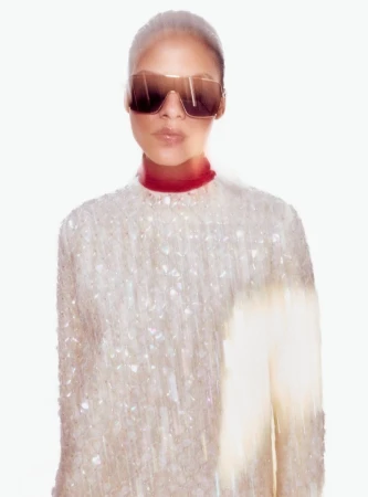 A stylish adult female is posing indoors while wearing sunglasses and a white sequin-covered top against a bright white background.