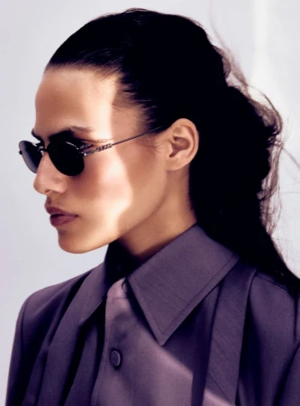 Elegant portrait of a woman with dark hair in stylish sunglasses and lavender coat, against a clean, bright white backdrop.