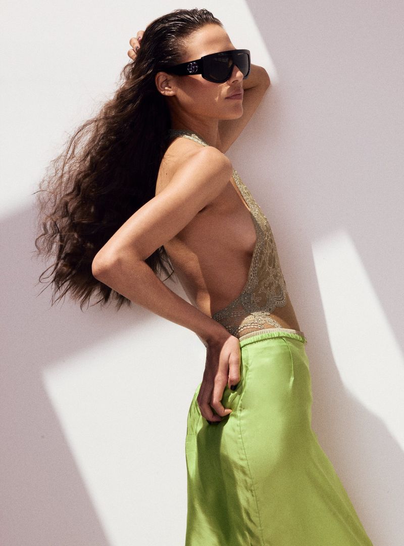 Brunette woman poses in a sleeveless beaded top and green skirt against a white wall, accessorized with large black sunglasses.
