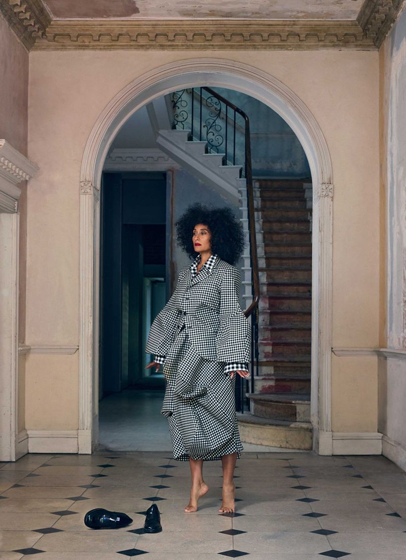 Woman with afro in black-and-white checkered outfit stands barefoot in hallway with archway and staircase, shoes discarded nearby.
