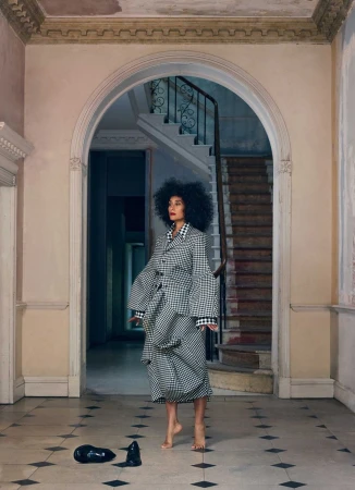 Woman with afro in black-and-white checkered outfit stands barefoot in hallway with archway and staircase, shoes discarded nearby.