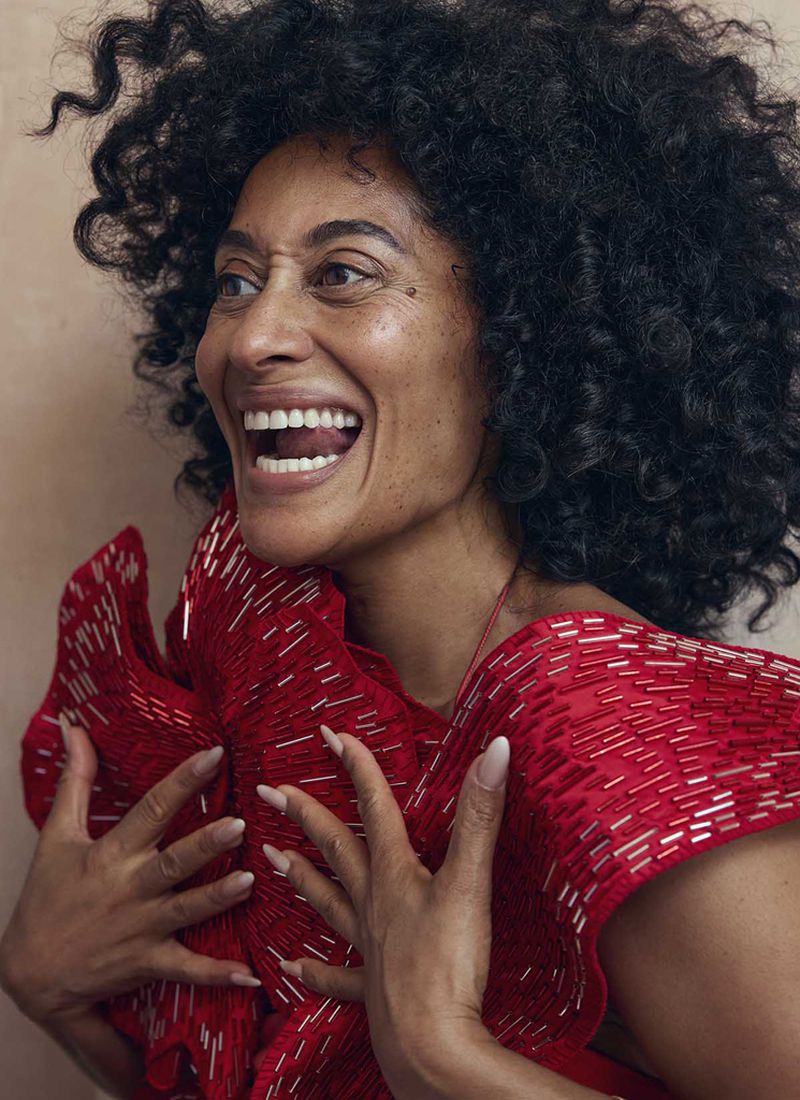 Portrait of Tracee Ellis Ross smiling, with curly hair and a vibrant red textured dress, hands clasped in front against a neutral backdrop.
