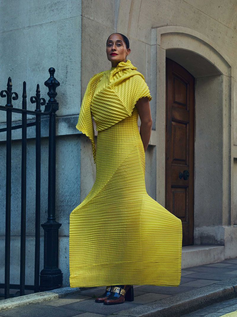 Adult woman poses in a pleated yellow dress in front of a stone wall and wrought iron fence near a wooden door on a city street.