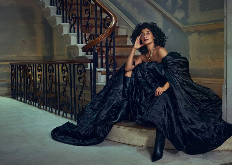 Adult female with curly dark hair sitting on a staircase wearing an off-the-shoulder black dress and high-heeled boots in an indoor setting.