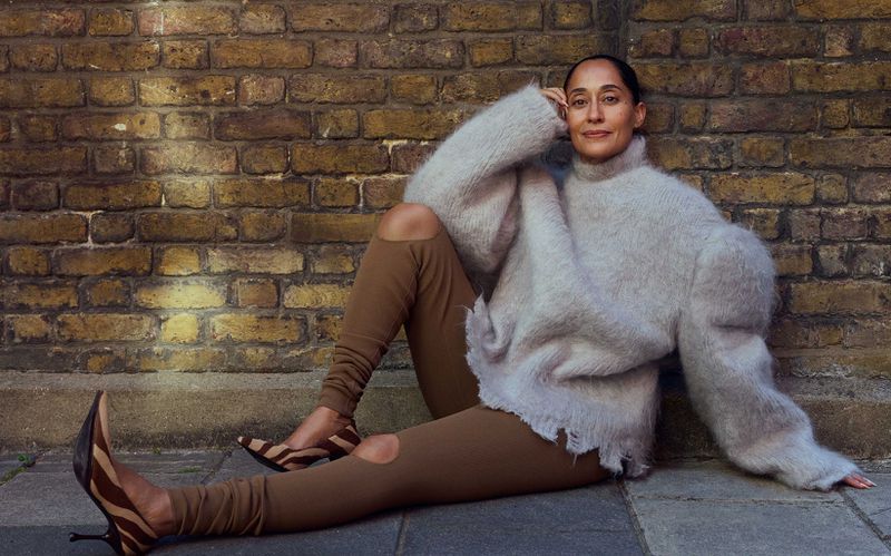 Adult woman in a beige sweater and brown leggings sitting against a brick wall and wearing zebra-print heels poses in an urban setting.